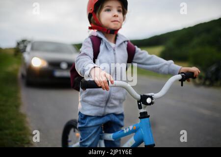 Ritratto di piccola ragazza eccitata in bicicletta su strada con auto dietro di lei, concetto di educazione sulla sicurezza stradale. Foto Stock