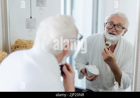 L'uomo anziano rasa la barba in bagno, guardando nello specchio. Concetto di routine mattutina. Foto Stock
