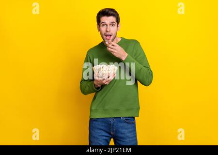 Foto di positivo impressionato giovane uomo indossare maglione verde mangiare gustoso popcorn isolato sfondo di colore giallo Foto Stock