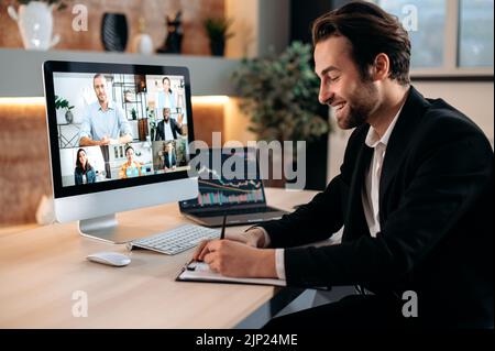 Positivo successo uomo d'affari caucasico in tuta, sedersi in un posto di lavoro, negoziare con i partner tramite videochiamata, discutere di nuovo progetto, termini di cooperazione, prende appunti. Videoconferenza in ufficio. Foto Stock