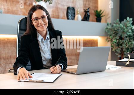 Ritratto di fiduciosa donna caucasica broker in ufficio. Bella donna bruna intelligente di successo, top manager aziendale o trader, utilizza il laptop, documenti di studio, guarda la fotocamera, sorridendo Foto Stock