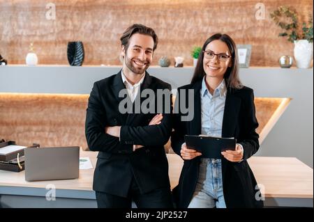 Ritratto di due dipendenti di successo caucasici fiduciosi, colleghi di lavoro, un uomo e una donna, in piedi vicino al desktop in un ufficio moderno, guardare la fotocamera, sorridere amichevole Foto Stock