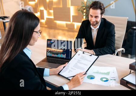 Concetto di cooperazione. Specialista di assunzione in una società di negoziazione, siede su una scrivania di fronte a un computer portatile, conduce un colloquio di assunzione con un uomo caucasico intelligente, concentrato a studiare il curriculum del candidato Foto Stock