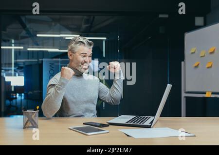 Riuscito e felice proprietario senior grigio-haired investitore maturo uomo d'affari che lavora in ufficio moderno utilizzando il laptop, l'uomo che celebra la vittoria trionfo, tenendo le mani in su e sorridendo, gesto di successo Foto Stock
