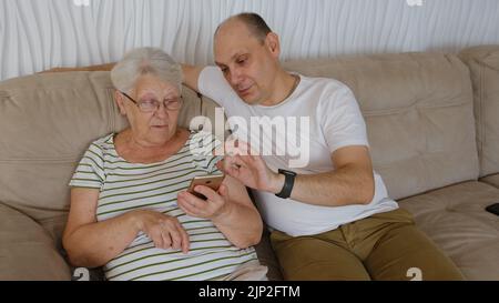 Nel soggiorno su un lato del divano seduta anziana 80s nonna con adulto 40s figlio, donna con smartphone mostra al vecchio uomo nuova app, teac Foto Stock