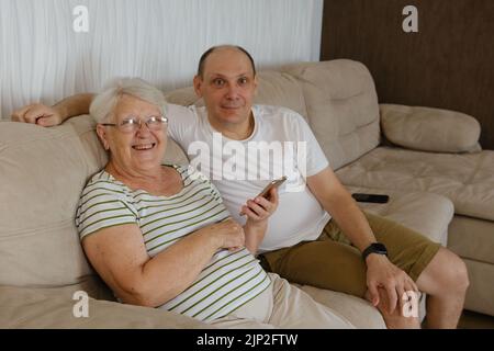 Nel soggiorno su un lato del divano seduta anziana 80s nonna con adulto 40s figlio, donna con smartphone mostra al vecchio uomo nuova app, teac Foto Stock