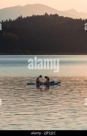 Due persone sedute su una Stand-Up-Paddle nel mezzo di un lago panoramico e ammirate il caldo tramonto Foto Stock