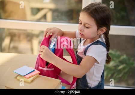 Deliziosa studentessa europea mette un notebook in zaino rosa, preparandosi per il primo grado di scuola elementare Foto Stock