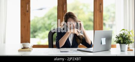 Donna d'affari asiatica sorridente al lavoro che parla al telefono e prende appunti, seduto al suo posto di lavoro in ufficio, spazio copia. Banner. Foto Stock