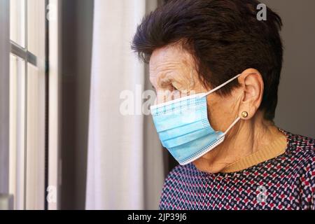Donna anziana con una maschera all'interno della sua casa che guarda fuori dalla finestra con paura dell'esterno, a causa del coronavirus Covid-19. Foto Stock