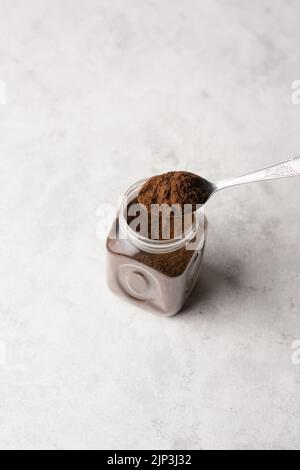 caffè macinato in un cucchiaio di metallo sulla parte superiore del vaso di vetro, posto su una superficie bianca testurizzata, sfondo morbido a fuoco con spazio per le copie Foto Stock