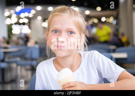 Ritratto di una ragazza un po 'affamata divertente mangiare freddo delizioso gelato in una tazza di waffle seduti in un caffè, sfondo scuro, bokeh. Un bambino gode di ghiaccio c Foto Stock