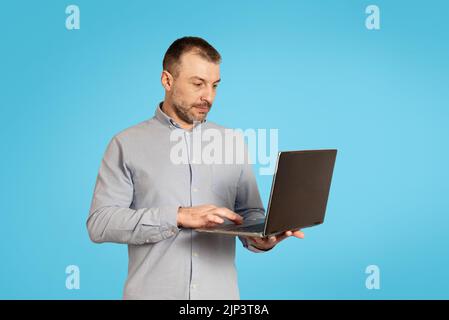Uomo d'affari di mezza età che usa il computer portatile che lavora in linea, sfondo blu Foto Stock