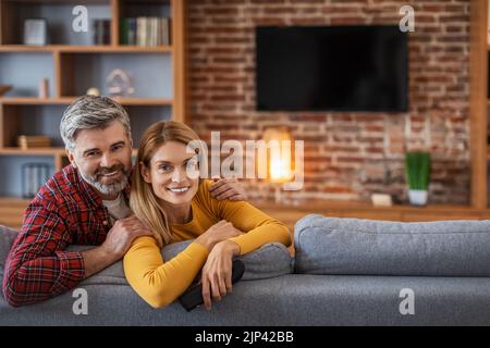 Felice uomo e donna europea adulti guardando la macchina fotografica, godere di relax nel soggiorno interno con tv con schermo vuoto Foto Stock
