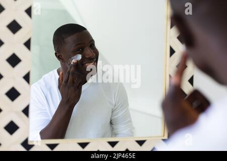 Uomo afroamericano sorridente che applica il moisturizer di faccia che osserva nello specchio della stanza da bagno Foto Stock