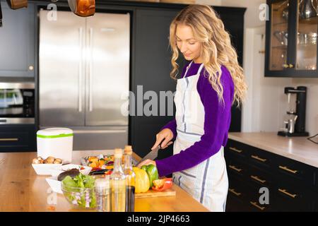 Donna caucasica felice in piedi in cucina con grembiule che trita verdure Foto Stock