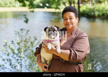 Vita in su ritratto di donna matura nera che tiene carino pug cane e guardando la macchina fotografica in bellissimo parco scenario, copia spazio Foto Stock