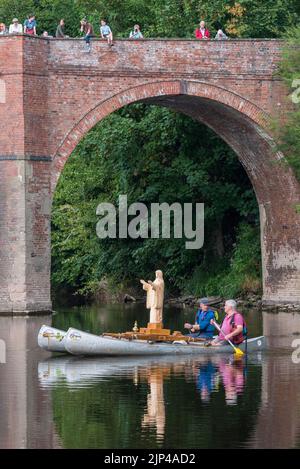 Il viaggio di nostra Signora delle acque e della Wye. Una statua in legno appena scolpita della Vergine Maria è stata fatta galleggiare 75 miglia lungo il fiume Wye, da Hay-on-Wye a Monmouth, dal 15th - 19th agosto 2022. Lo scultore Philip Chatfield (in rosa) e un amico, Callum Bulmer (in blu), stanno facendo il viaggio per sensibilizzare gli alti livelli di inquinamento nel Wye, uno dei fiumi più importanti e belli della Gran Bretagna. Sono visti qui passando sotto il vecchio ponte a Bredwardine, Herefordshire. Foto Stock