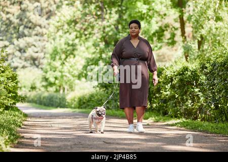 Ritratto completo di elegante donna matura vestito mentre cammina cane piccolo nel parco, spazio copia Foto Stock