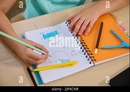 Vista dall'alto di un copybook di matematica e di una mano di un scolaro che tiene una matita per risolvere i problemi in una lezione di geometria online Foto Stock
