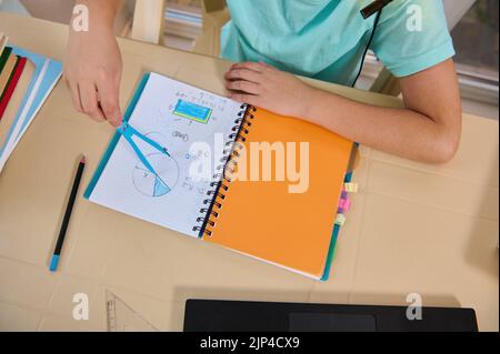 Vista dall'alto della mano di uno scolaro che tiene la bussola, disegnando un cerchio mentre risolve i problemi in una lezione di geometria online Foto Stock