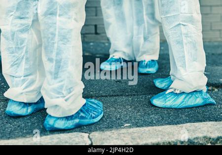 Gambe e piedi degli operatori sanitari che indossano indumenti protettivi o tute in materiale hazmat all'aperto durante la pulizia. Primo piano di un team di professionisti medici Foto Stock