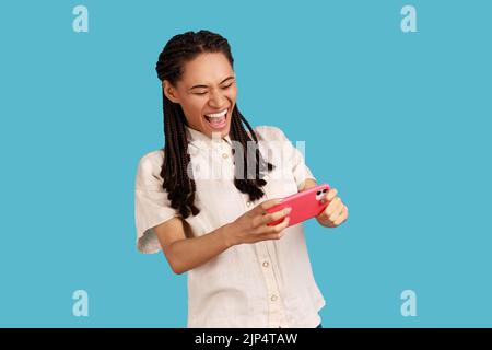 Donna emozionale felice con i greggi neri fissa la fotocamera dello smartphone, gioca, urla felicemente mentre il gioco sorride eccitato, indossando una camicia bianca. Studio in interni isolato su sfondo blu. Foto Stock