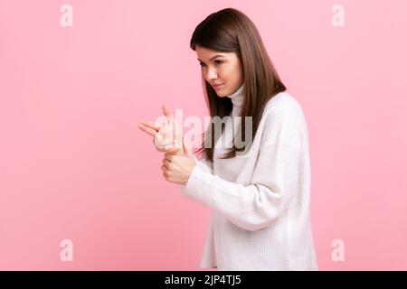 Donna criminale pericolosa che punta la pistola, minacciando di uccidere, sparando con gesto d'arma, indossando un maglione bianco casual. Studio in interni isolato su sfondo rosa. Foto Stock