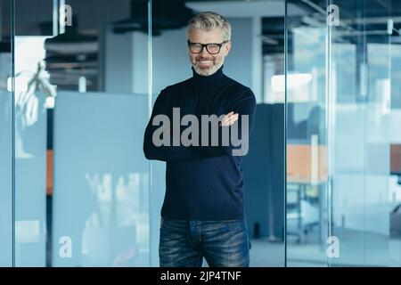 Ritratto di successo finanziatore maschio, uomo d'affari maturo sorridente e guardando la macchina fotografica con braccia incrociate, uomo con capelli grigi che lavora in ufficio, capo che indossa occhiali e jeans Foto Stock