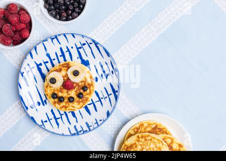 Frittelle americane decorate come un sorriso e facce felici con lamponi, mirtilli e banana. Cibo per bambini, allegro e creativo. Vista dall'alto. Foto Stock