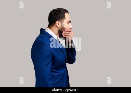 Vista laterale di attraente uomo d'affari bearded che chiude la bocca con la mano, ha paura di dire troppo, imbarazzo, indossando il vestito ufficiale di stile. Studio in interni isolato su sfondo grigio. Foto Stock