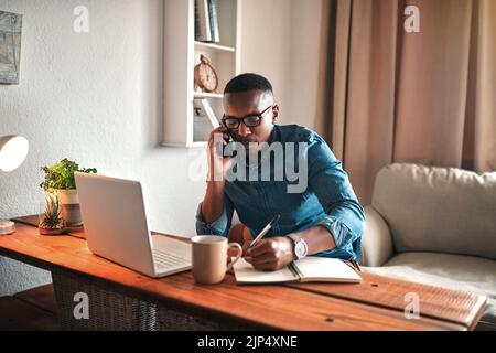 Giovane imprenditore al telefono mentre prende appunti alla sua scrivania. Uomo professionista che lavora lontano da casa con la tecnologia. Il maschio afro-americano ha un Foto Stock