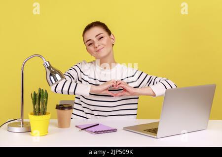 Ritratto di soddisfatta bella donna con sorriso gentile seduta sul posto di lavoro, mostrando forma romantica del cuore con la mano, esprimendo l'amore. Studio interno girato isolato su sfondo giallo. Foto Stock