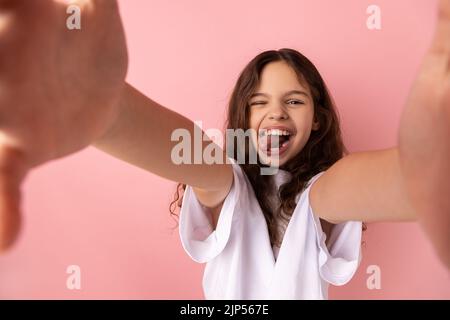 Ritratto di divertente bambina che indossa una T-shirt bianca con un'espressione facciale positiva che prende selfie e mostra la lingua fuori, punto di vista della foto. Studio in interni isolato su sfondo rosa. Foto Stock