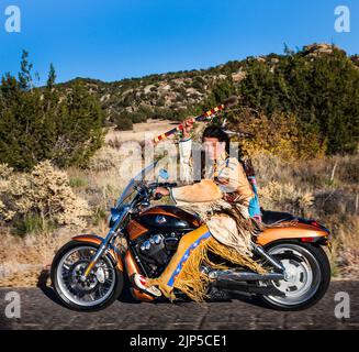 L'uomo indiano americano tradizionalmente vestito tiene in su il bastone del colpo mentre guida una motocicletta di Harley Davidson giù l'autostrada di paese Foto Stock