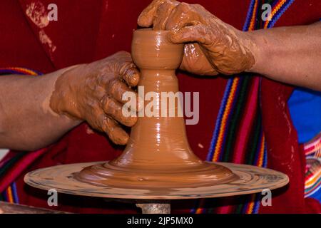 Primo piano mani ricoperte di fango di un adulto ecuadoriano che prepara una tazza di argilla sulla ruota di un vasaio. Uomo vestito con poncho tradizionale Foto Stock