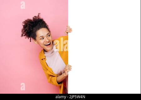 Stupito africano americano giovane donna riccio, peeking fuori da lavagna bianca con modello mockup e spazio di copia per la presentazione o la pubblicità e guarda esso, stand su sfondo rosa, sorrisi Foto Stock