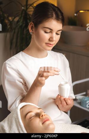 Cosmetologo preparazione maschera cosmetica idratante per applicarla al viso del paziente. Donna che applica la crema per il viso. Concetto spa. Foto Stock