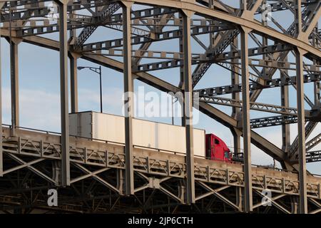 Un camion di trasporto rosso con un rimorchio bianco è visto viaggiare attraverso un ponte a traliccio. Foto Stock