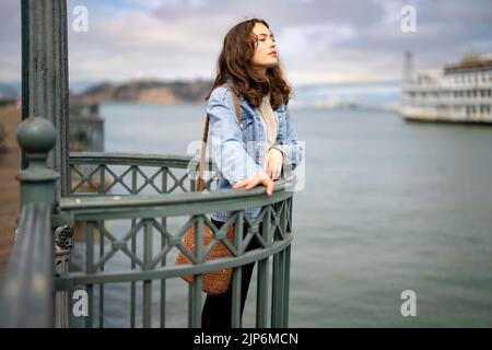 Ritratto senza posa della giovane donna pensierosa sul molo che si affaccia sulla baia di San Francisco Foto Stock