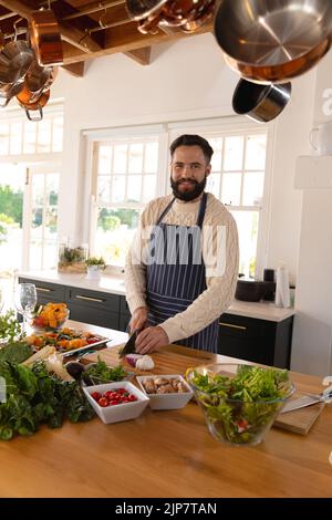 Ritratto di felice uomo caucasico con barba tritare verdure con grembiule in cucina Foto Stock