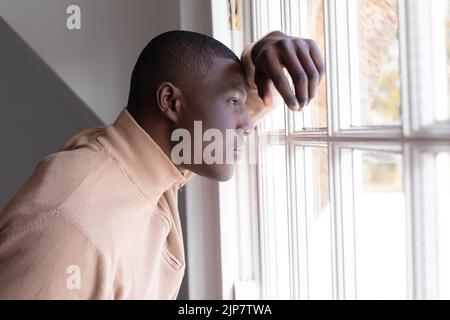 Uomo afro-americano premuroso a casa che si appoggia e guarda fuori dalla finestra Foto Stock