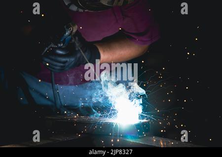 Un uomo che esegue la saldatura e la smerigliatura sul posto di lavoro in officina, mentre le scintille 'volano' tutto intorno a lui. Indossa un casco di protezione Foto Stock