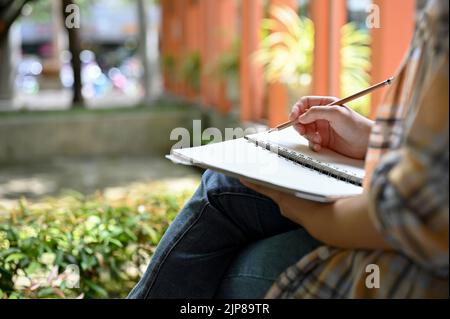 Una ragazza freelancer hippster notando le sue idee creative in un notebook mentre rilassarsi siede nel parco della città. Immagine ritagliata Foto Stock
