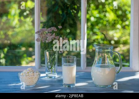 Latte in una brocca in un bicchiere e formaggio casolare sul davanzale vicino alla finestra della casa in una giornata estiva vicino al giardino, primo piano. Lotti di prod. Caseario Foto Stock