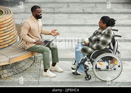 Scatto completo di coppia nera adulta con donna in sedia a rotelle che chiacchiera all'aperto in città e si diverte a bere un caffè Foto Stock