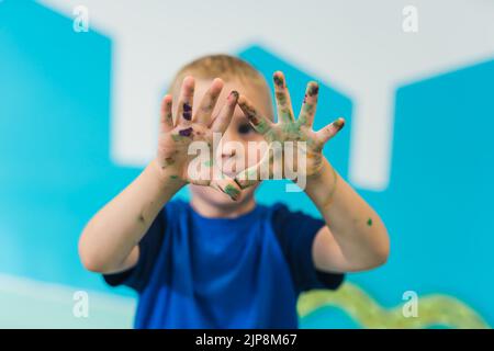 Scuola materna. Ragazzo biondo carino che mostra le sue palme colorate dipinte. Classe d'arte con pittura a dita per bambini abilità sensoriali, creatività e sviluppo di immaginazione Foto Stock