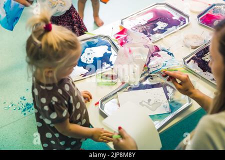 Piccola ragazza caucasica carina con altri bambini pittura latte, aggiungendo colorante alimentare al latte con un insegnante in classe presso la scuola materna. Foto di alta qualità Foto Stock