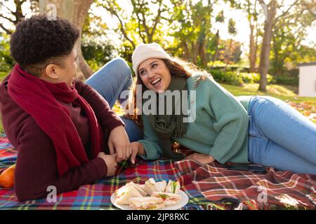 Felice coppia biraciale sdraiato su un tappeto con pic-nic e parlare nel giardino d'autunno Foto Stock