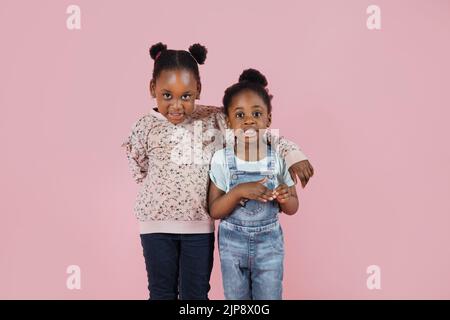 Due felici bambini divertenti, ridendo piccole ragazze africane, in abiti pastello casual, in posa su sfondo rosa parete. Due suore per bambini si abbracciano l'una con l'altra Foto Stock